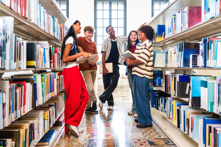 Junge Studierende lächeln und arbeiten zusammen, während sie in einer hellen Universitätsbibliothek in den Bücherregalen recherchieren und gemeinsam lernen.